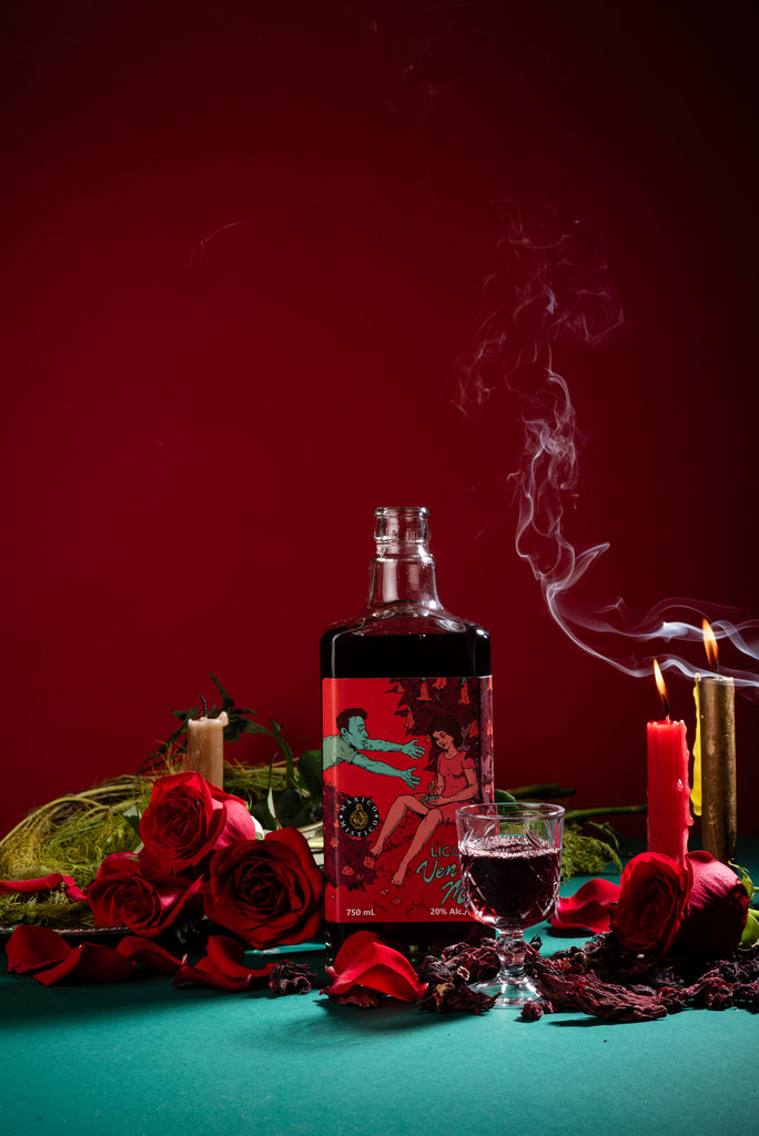 Bottle of Ven a mi liquor, candles, and red flowers on a green surface with red background. Moodshot Ven a mi